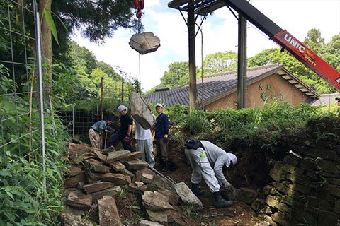 シカが石垣の上を走って崩してしまった部分の修復。講習会も兼ねた今回（2017年7月22日実施）は、重機も使って慎重に行われました。あうんの呼吸でテキパキと作業が進み、チームワークも抜群です